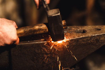 The blacksmith manually forging the red-hot metal on the anvil in smithy with spark fireworks.