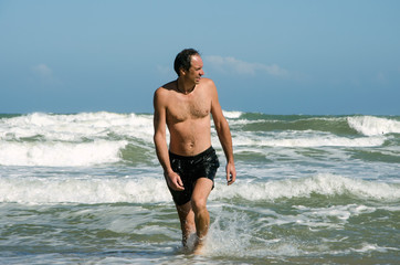 Fototapeta na wymiar A man in swimming trunks comes out of the water onto the shore, looks away and smiles. In the background are powerful waves of the surf. Clear sky, sunny day.
