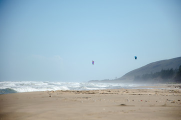 Sea beach with strong surf. Rocky cape, strong wind bends the trees and spreads a spray of waves. In the background are the kitesurfing sails in the sky. Sunny day.