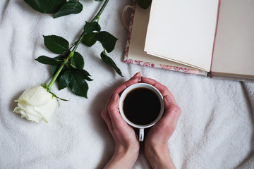 Woman's hands holding a cup of coffee on the white bedspread with white rose and notebook on it.