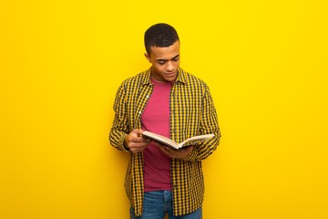 Young afro american man on yellow background holding a book and enjoying reading