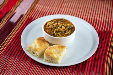 A bowl of hearty soup with chunks of vegetables and grains, served with pieces of crusty bread on a white plate, accompanied by a wooden utensil, set against a vibrant red and gold striped background.