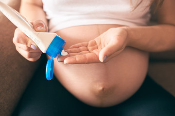 Selective focus of female hands keeping cream and applying it on big belly. Young pregnant woman sitting on sofa and caring about skin. Concept of expectation and parental love.