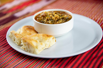 A bowl of hearty soup with chunks of vegetables and grains, served with pieces of crusty bread on a white plate, accompanied by a wooden utensil, set against a vibrant red and gold striped background.