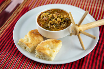 A bowl of hearty soup with chunks of vegetables and grains, served with pieces of crusty bread on a white plate, accompanied by a wooden utensil, set against a vibrant red and gold striped background.