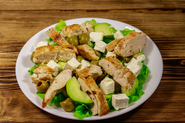 Tasty salad with fried chicken breast, green olives, feta cheese, avocado, lettuce and olive oil on wooden table