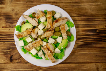 Tasty salad with fried chicken breast, green olives, feta cheese, avocado, lettuce and olive oil on wooden table. Top view