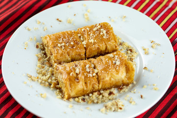 Two pieces of golden, flaky baklava on a white plate, garnished with crushed nuts, set against a vibrant red and gold striped background.