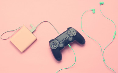 Gamepad is charging from the power bank, earphones on pink background, Modern devices, top view..