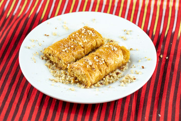 Two pieces of golden, flaky baklava on a white plate, garnished with crushed nuts, set against a vibrant red and gold striped background.