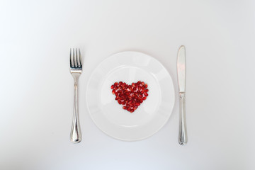 On a white plate from pomegranate seeds composite heart shape. Knife and fork next to the plate. White background. Flat layer.