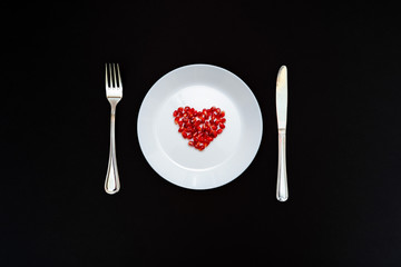 On a white plate from pomegranate seeds composite heart shape. Knife and fork next to the plate. Black background. Flat layer.