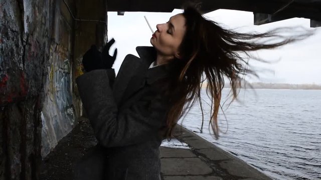A young woman in a gray coat smokes near the river.