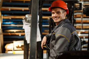 Fototapeta premium Young smiling worker wearing orange helmet and gray uniform sitting on forklift and transporting hardware on metal stock. Happy loader turning back, rutting arm on steering wheel and posing at camera.