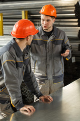 Two workers wearing uniforms and orange hardhats standing together and talking emotionally at metal warehouse. Male leaning on metal sheet and listening to his coworker explaining and gesturing.