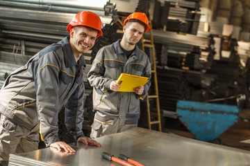 Side view of two workers of metal stock in gray uniforms and orange hardhats operating together...