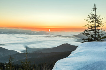 The fantastic scenery of the winter Carpathians with a snow texture in the foreground with dense fog in the mountain valley and the morning sun on the horizon