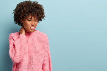 Dissatsfied woman keeps hand on neck, clenches teeth in displeasure, has sad expression, wears oversized pink sweater, feels stressful and sick, isolated over blue background with blank space aside