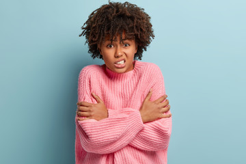Sad mixed race young woman clatters teeth, crosses hands over chest, shakes from cold weather, wears pink oversized jumper, isolated over blue background. Winter time and frosty day concept.