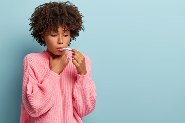 Sad ill young Afro American girl keeps thermometer in mouth, measures temperature, wears oversized pink jumper, has thore throat, cold or flu, models over blue background with empty space aside