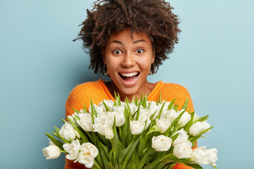 Photo of pleasant looking African American woman enjoys pleasant scent of beautiful flowers, laughs...