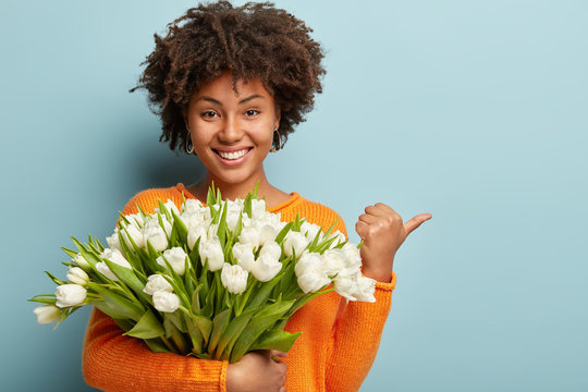 Beautiful pleasant looking cheerful woman with calm happy expression, holds bouquet of white tulips in front of chest, has natural beauty, points with thumb, shows free space for your promotion