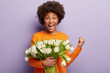 Waist up shot of joyful woman raises fist in triumph, enjoys date with guy, recieves white tulips, wears orange jumper, isolated over purple background. Positive dark skinned lady with flowers bouquet