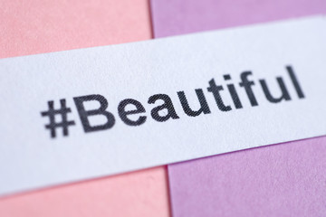 Popular hashtag 'beautiful' printed on white sheet of paper on a pink and purple background. Close up.