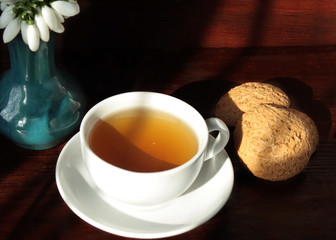 White cup on a saucer with oatmeal cookies near a vase with snowdrops on the table