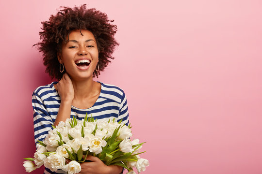 Emotive pleased black woman expresses happiness, wears sailor casual jumper, smells scent of white tulips, poses against pink background with blank space for your promotional content enjoys spring day