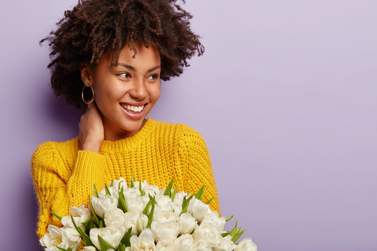 Sideways shot of pleasant looking young woman with crisp hair, toothy smile, wears knitted yellow sweater, holds white flowers recieved on Womens Day, isolated over purple wall, blank space aside