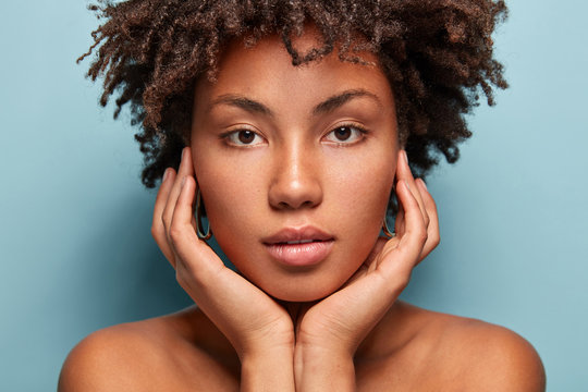 Close up shot of black young woman with Afro hairstyle, touches cheeks with both hands, has bare shoulders, tender look isolated over blue background, looks directly at camera. Horizontal shot