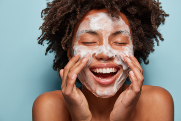 Spa, therapy and beauty concept. Satisfied black woman washes face with white soap, has cleansing foam, purification of face, cleans pores, shows bare shoulders, isolated over blue studio wall - obrazy, fototapety, plakaty