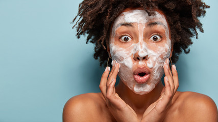 Cropped image of overwhelmed dark skinned lady has soap on face, touches cheeks, surprised...