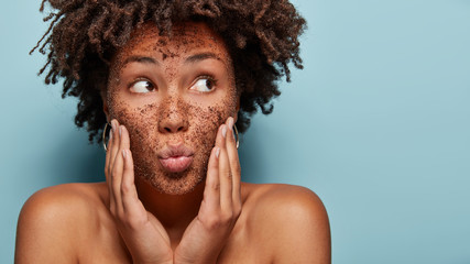 Curious black lady focused aside has coffe scrub on face applies mask on face, focused aside, has bare shoulders, isolated over blue background with blank space for promotion. Body care. Cosmetology - obrazy, fototapety, plakaty