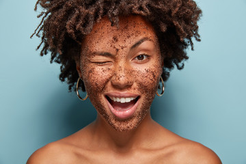 Image of pleased young curly woman blinks eye, has positive expression, applies black coffee mask, does peeling of skin, poses with bare shoulder, isolated over blue background, visits spa salon
