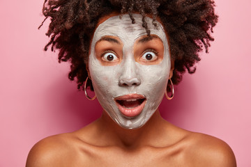 Surprised young woman with shocked expression, applies facial clay mask, has healthy natural skin, amazed by effect of beauty cream, wears earrings, isolated over pink background. Dry complexion