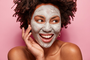 Photo of glad black woman has clay mask, enjoys facial treatments, keeps gaze aside, touches face, has spa therapy, broad smile with white teeth, isolated over pink background. Perfect skin concept