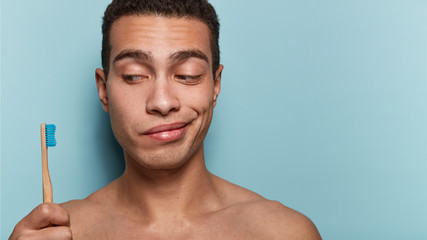 Mouth hygiene concept. Photo of attractive male model holds new toothbrush, ready to brush teeth, has morning bathroom routine, poses naked against blue background with blank space for information