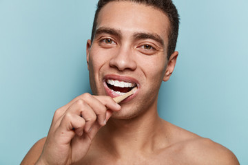 Smiling young man with healthy teeth, holds toothbrush, cares of dental hygiene, looks pleased, has dark eyes, short hairstyle, muscular body, isolated over blue studio wall. Daily routines.