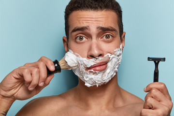 Men, grooming and shaving concept. Puzzled Caucasian man going to shave beard, holds sharp razor and brush, poses half naked, busy with daily routine in morning, isolated over blue background.