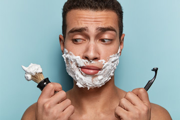 Macho man with naked torso has shaving gel on face, frustrated facial expression, holds brush and...