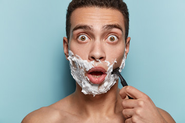 Close up shot of surprised emotive stunned man with eyes bugged, shaves bristle with sharp razor, uses shaving gel, poses with naked torso, keeps mouth opened, isolated over blue background.