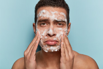Image of displeased European man washes face with soap, has discontent expression because of acnes,...