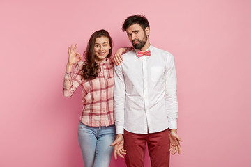 Horizontal shot of cheerful woman with okay gesture, gives approval, agrees to work together with groupmate who stands clueless, spreads hands, wears elegant clothes, isolated over pink studio wall