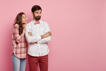 Thoughtful bearded young man keeps arms folded, wears white shirt with bowtie, being deep in thoughts, beautiful woman in checkered shirt stands closely, keeps lips folded, wants to support.