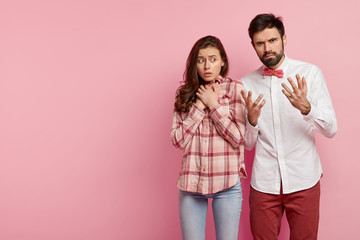Photo of annoyed bearded young man gestures in puzzlement, worried lady keeps hands on chest, face problems, stand next to each other, isolated over rosy background with empty space on left side