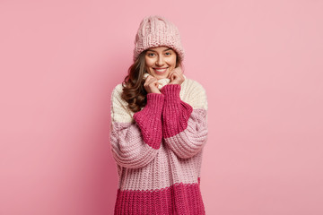 Portrait of happy young European woman has gentle smile, keeps hands under chin, looks directly at camera with satisfied expression, wears knitted winter hat and loose sweater, enjoys spare time