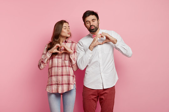 Love symbol. Affectionate romantic woman and man keep lips folded, make heart gesture, express good feelings, wear shirt and trousers, want to kiss each other, isolated over pink background.