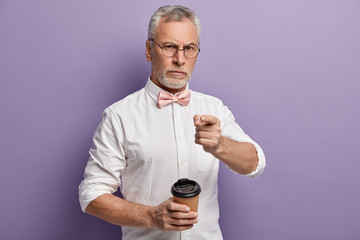 Confident mature wrinkled unshaven man in eyewear, wears elegant white shirt, pink bowtie, holds takeaway coffee, points at camera with self confident expression, isolated over purple background